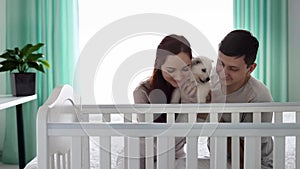 A young married couple with a small white puppy are looking into the crib where the newborn is sleeping. The concept of