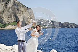 Young married couple uncorking champagne by the sea after their wedding