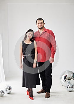 Young married couple, wearing red and black fancy clothes, standing in light room near big window. New Year party celebration.