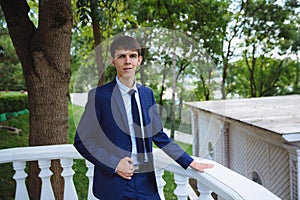 A young married man in a strict blue suit is posing on a large balcony overlooking the park on her wedding day. An