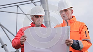 Young and mature architect with hard hat and protective clothing discussing a plan on a construction site