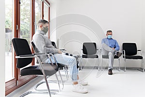 Young and mature man with face masks sitting in a waiting room of a hospital or office