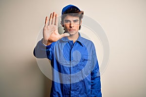 Young mechanic man wearing blue cap and uniform standing over isolated white background doing stop sing with palm of the hand