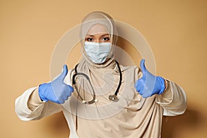 Young medic wearing hijab and medical mask showing thumbs up gesturing with hands in medical protective gloves
