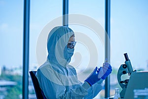 Young medical scientist wearing protective white jumpsuit and face mask with glasses testing sample with microscope in clinical