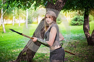 Young medieval woman warrior dressed in chain mail armor with a spear in her hands on the background of the forest.