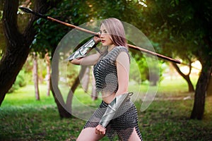 Young medieval woman warrior dressed in chain mail armor with a spear in her hands on the background of the forest.