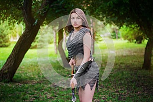 Young medieval woman warrior dressed in chain mail armor with a spear in her hands on the background of the forest.