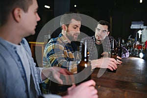 Young men in casual clothes are talking, laughing and drinking while sitting at bar counter in pub