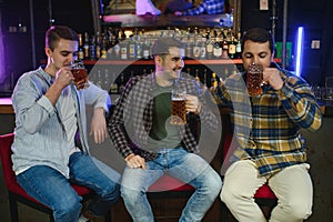 Young men in casual clothes are talking, laughing and drinking while sitting at bar counter in pub