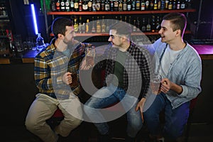 Young men in casual clothes are talking, laughing and drinking while sitting at bar counter in pub