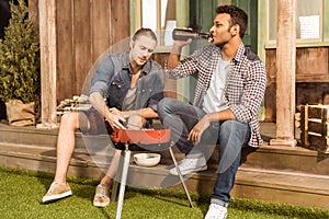 Young men drinking beer and preparing meat on outdoor grill