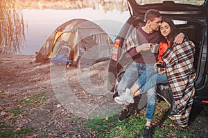 Young man embrace woman. They sit in trunk. Model is covered with blanket. Couple is at lake. There is tent at waterline.