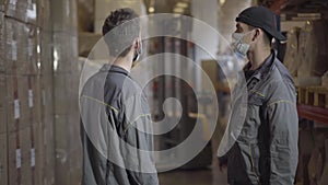 Young men in face masks standing in warehouse and talking. Professional experts discussing storage of finished