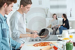 Young men making pizza girlfriends sat on sofa