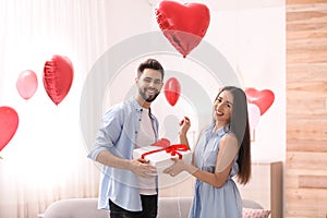 Young man presenting gift to his girlfriend in room decorated with heart shaped balloons. Valentine`s day celebration