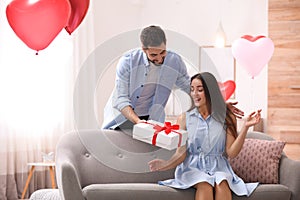 Young man presenting gift to his girlfriend in room decorated with heart shaped balloons. Valentine`s day celebration