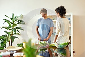 Young man smiling, cutting vegetables while woman talking and watching him. Vegetarians preparing healthy meal in the
