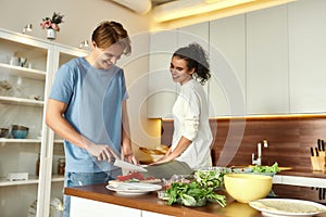 Young man smiling, cutting vegetables while woman talking and watching him. Vegetarians preparing healthy meal in the