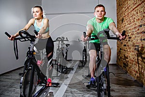 Young man and woman biking in the gym, exercising legs doing cardio workout cycling bikes.