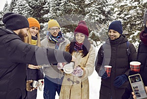 Young men and women with hot tea in paper disposable cups walk through snowy forest on winter day.