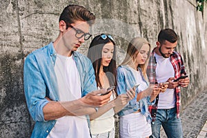 Young men and women are standing on grey background and looking at their phones. They are serious and concentrated.