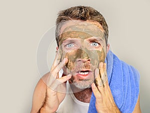 Young messy funny man looking at himself horrified in bathroom mirror with green cream on his face applying beauty facial mask pro