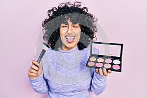 Young middle east woman holding makeup brush and blush sticking tongue out happy with funny expression