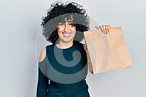 Young middle east woman holding take away paper bag looking positive and happy standing and smiling with a confident smile showing