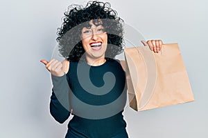 Young middle east woman holding take away paper bag pointing thumb up to the side smiling happy with open mouth