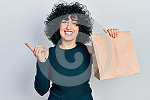 Young middle east woman holding take away paper bag smiling happy pointing with hand and finger to the side