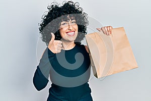 Young middle east woman holding take away paper bag smiling happy and positive, thumb up doing excellent and approval sign