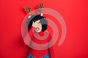 Young middle east woman wearing cute christmas reindeer horns peeking in shock covering face and eyes with hand, looking through