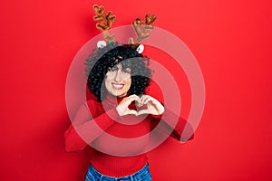 Young middle east woman wearing cute christmas reindeer horns smiling in love doing heart symbol shape with hands