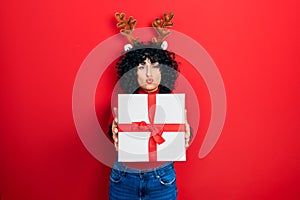 Young middle east woman wearing deer christmas hat holding gift looking at the camera blowing a kiss being lovely and sexy