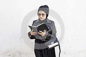 Young middle-eastern student girl holding in hands stack of notepads and her handbag. Modern Muslim woman wearing traditional