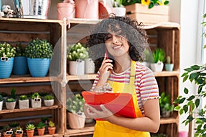 Young middle eastern woman florist talking on smartphone reading clipboard at florist