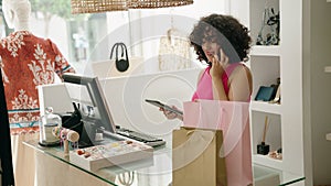 Young middle eastern woman shop assistant using touchpad talking on smartphone at clothing store