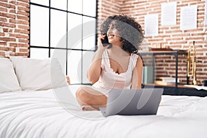Young middle eastern woman using laptop talking on smartphone at bedroom