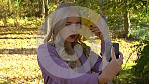 Young millenial attractive blonde girl with autumn leaf making selfie on her mobile smartphone in autumn forest park at