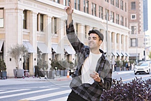 Young Millennial Male Raises His Hand to Signal an Uber Driver - Rideshare Request Using Cell Phone in His Hand - During the Day w