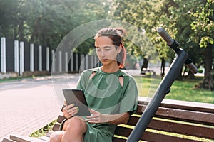 Young millennial woman uses a tablet while sitting on a bench.