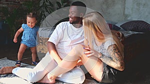 Young mixed culture family couple relaxing on carpet watching little daughter.