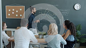 Young mixed race man making report pointing at chalkboard and talking to milti-ethnic group of colleagues