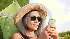 Young Mixed Race Tourist Girl in Sunglasses and Straw Hat Using Mobile Phone in Hammock in Park at Natural Sunset