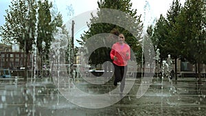 Young mixed race woman running in the rain near city park fountains, slow motion shot