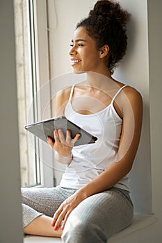 Young mixed race woman with tablet pc looking out the window, feeling cozy while sitting alone by the window at home