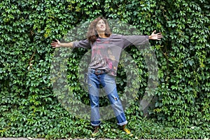 A young model beautiful woman girl in denim trousers stands near a wall overgrown with climbing plants. Background with plant