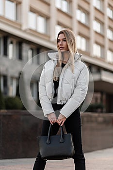 Young model of a beautiful woman with stylish black bag in a fashionable white jacket in jeans stands outdoors on an autumn day