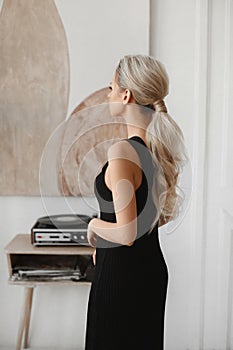 A young model woman with blond hair wearing a black evening dress posing in a minimalist interior